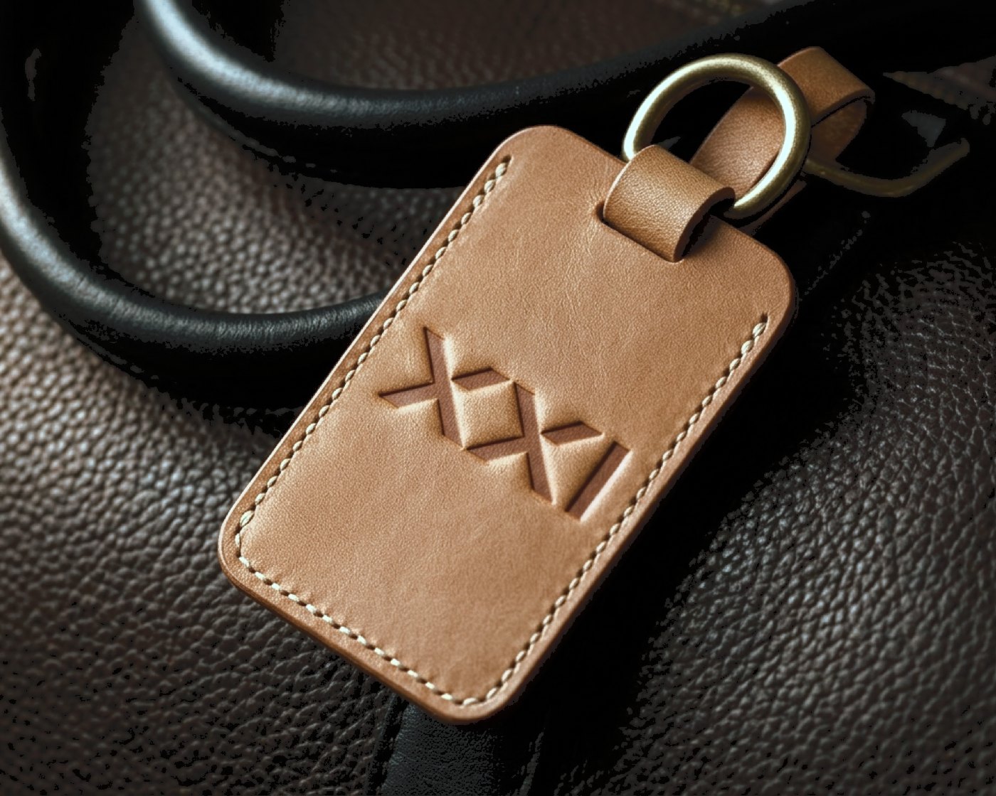 A tan leather luggage tag debossed with the XXI mark, clipped to a dark leather bag.