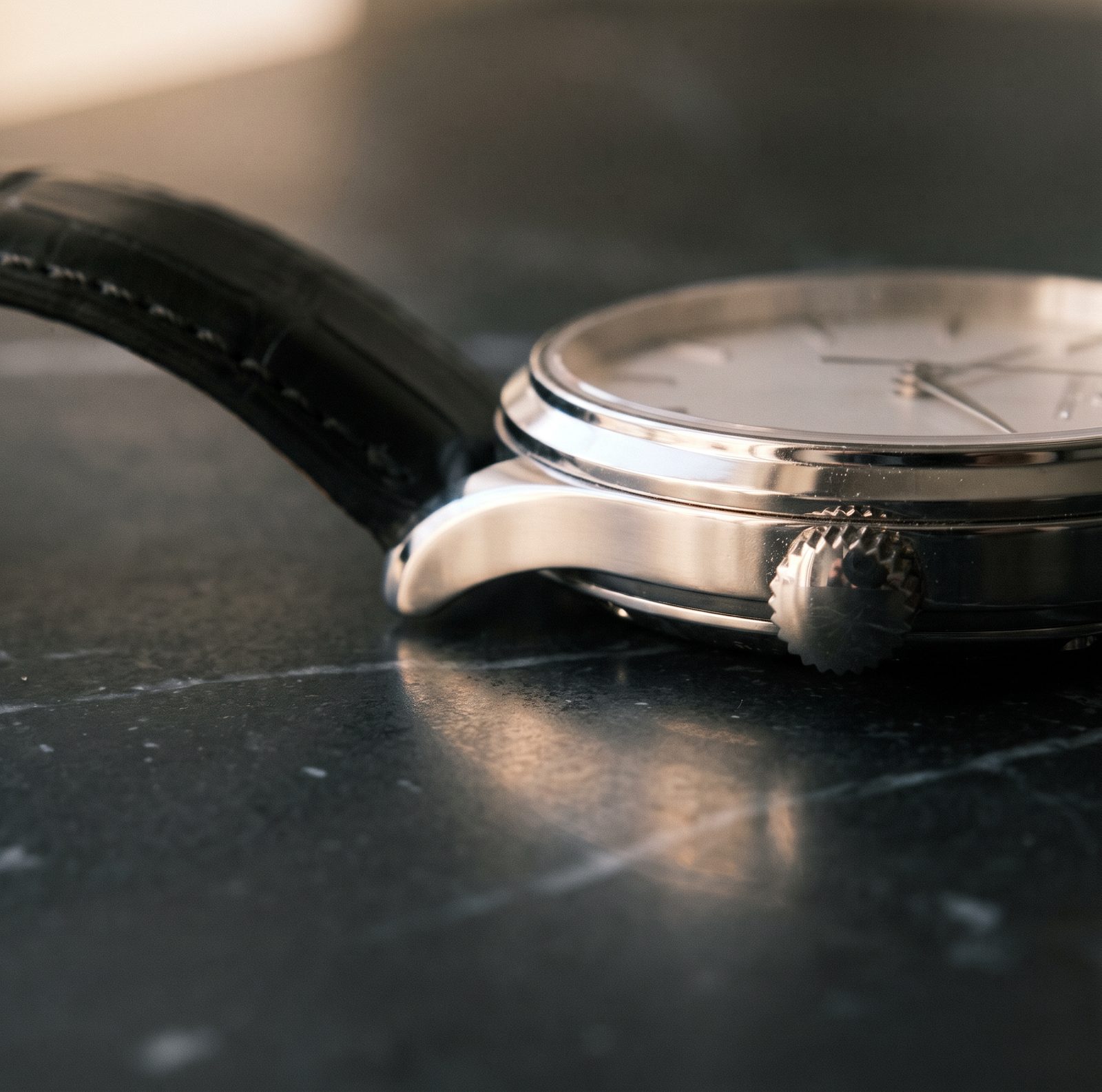 A steel wristwatch with a black leather strap lying on its side on dark marble.